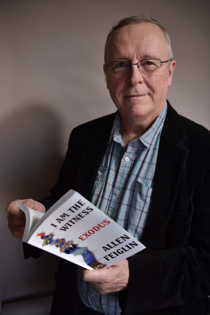 66-year-old man in button down shirt and blazer, holding an open book. The cover says "I am the Witness: Exodus."